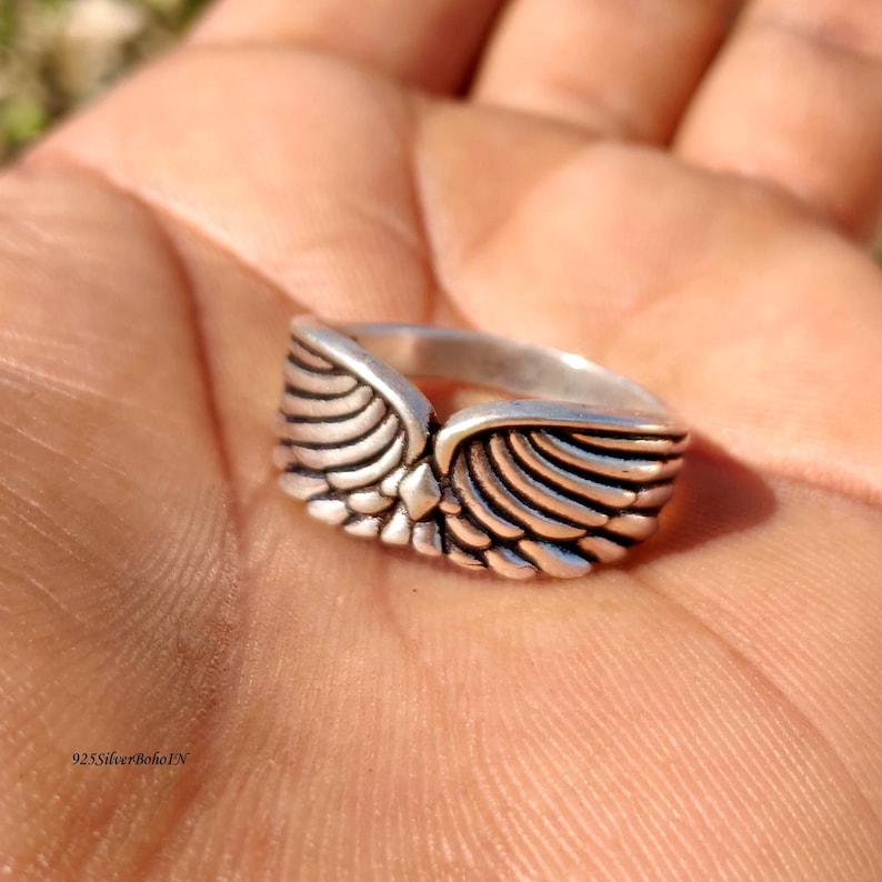 Pu&ograve; includere: Un anello d'argento con un design ad ali dettagliato. L'anello presenta un punto centrale dove le ali si incontrano, con intricati dettagli di piume. L'anello &egrave; mostrato in un primo piano, evidenziando l'artigianato e il design.