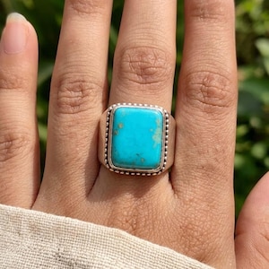 May include: A silver ring featuring a rectangular, bright turquoise stone with natural brown matrix inclusions. The stone is set in a smooth bezel with a decorative beaded border along the edge of the setting, mounted on a simple silver band.