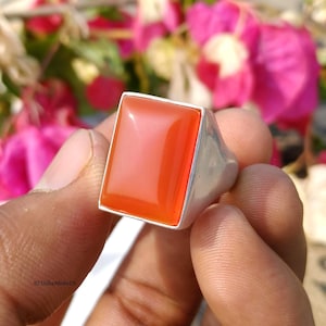 Peut inclure: Une bague en argent avec une grosse pierre précieuse orange rectangulaire. La monture de la bague est épaisse et polie, et la pierre précieuse a une surface lisse et réfléchissante. La bague est présentée sur un fond de fleurs roses.