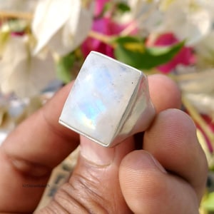 May include: A square-faced, silver-toned ring with a large, iridescent moonstone. The moonstone displays a play of color, with flashes of blue and yellow. The ring is held in a hand, with a blurred floral background.
