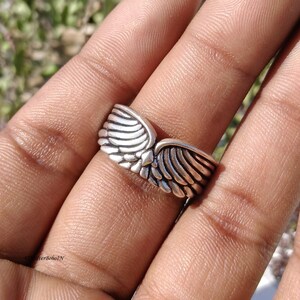 Pu&ograve; includere: Un anello color argento con un design ad ali. L'anello presenta due ali dettagliate che si incontrano al centro. Le ali hanno un aspetto strutturato e piumato e l'anello viene indossato su un dito.