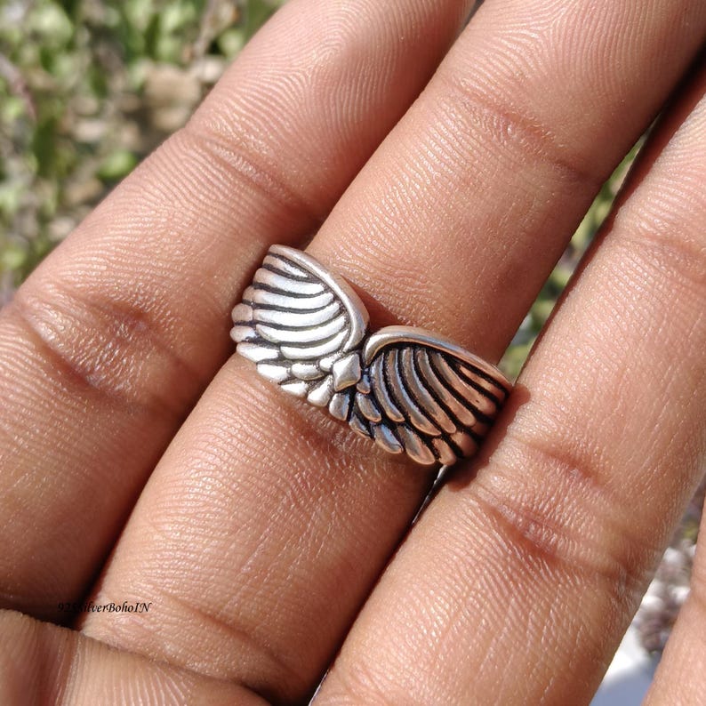 Pu&ograve; includere: Un anello color argento con un design ad ali. L'anello presenta due ali dettagliate che si incontrano al centro. Le ali hanno un aspetto strutturato e piumato e l'anello viene indossato su un dito.