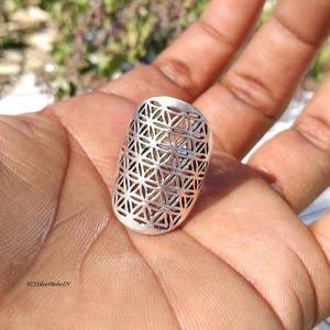 Pode incluir: Um anel prateado com um padrão geométrico de triângulos interligados. O anel tem uma forma oval alongada e é segurado em uma mão. O design é aberto, permitindo que a luz passe pelas aberturas triangulares. O texto "925SilverBohoIN" é visível.