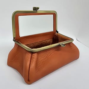 May include: A rectangular orange leather pouch with a textured surface and a brass-toned metal frame. The frame features a hinged opening mechanism with a central clasp and a clear rectangular window panel integrated into the top section.