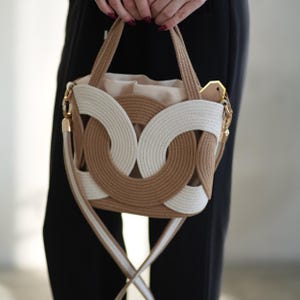 Op de afbeelding: Een beige en witte touwen handtas met een uniek lusontwerp. De tas heeft een beige binnenkant en goudkleurige hardware. Het heeft een schouderriem met witte en beige strepen en twee handvatten. De tas wordt vastgehouden door een persoon die een zwarte broek draagt.