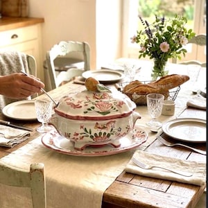 Peut inclure: Une table de salle à manger dressée pour un repas, avec une soupière à décor floral sur une assiette assortie. La table est garnie d'assiettes, de verres, de couverts et d'un panier à pain. Un vase de fleurs ajoute de la couleur.