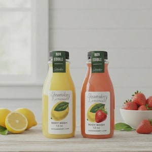 Könnte beinhalten: Zwei Flaschen Sheamaker Lemonade Duschgel, eine gelbe und eine rosa, mit schwarzen Verschlüssen und Etiketten. Die Etiketten zeigen eine Zitrone und eine Erdbeer-Illustration. Die Flaschen sind je 355 ml. Zitronen und Erdbeeren auf dem Holztisch.