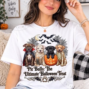 May include: A white t-shirt featuring a graphic of four pit bull puppies dressed for Halloween. One wears a vampire cape, one is a mummy, one sits in a pumpkin, and one has a skeleton print. Text reads: Pit Bulls: The Ultimate Halloween Treat.