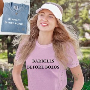 Könnte beinhalten: Ein hellblaues T-Shirt mit dem Text "BARBELLS BEFORE BOZOS" wird neben einer Frau gezeigt, die ein rosa T-Shirt mit demselben Text trägt. Die Frau trägt ein weißes Visier. Das T-Shirt hat einen Rundhalsausschnitt.