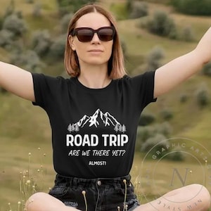 以下が含まれることがあります： 白い山のグラフィックと「ROAD TRIP ARE WE THERE YET？ALMOST！」という文字が入った黒いTシャツ。Tシャツは半袖で、丸いネックラインです。サングラスをかけています。