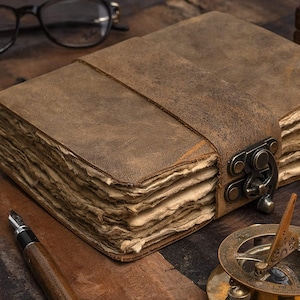 Peut inclure: Un journal en cuir marron avec une fermeture à boucle et une sangle. Le journal a des pages texturées et vieillies et une apparence rustique. Un stylo, des lunettes et une boussole sont également visibles.