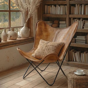 May include: A cozy reading nook featuring a brown leather butterfly chair with a patterned pillow. A wooden bookshelf is filled with books. The room has a large window with a view of trees and a wooden floor. Two small wicker side tables are present.