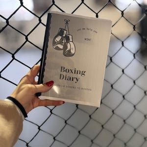 Puede incluir: Un cuaderno "Boxing Diary" de color plateado con encuadernación en espiral negra. La portada presenta un gráfico de dos guantes de boxeo y el texto "Let me help you WIN!" y "Boxing Diary FROM BOXERS TO BOXERS".