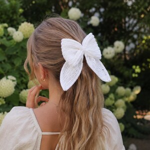 Könnte beinhalten: Ein großer, weißer Häkel-Haarschmuck in Form einer Schleife ist das Hauptmerkmal. Die Schleife hat ein detailliertes, strukturiertes Design. Der Hintergrund zeigt einen grünen Garten mit weißen Blüten, was eine weiche, natürliche Umgebung schafft.