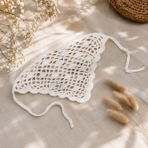 Könnte beinhalten: Ein weißes, offen gewebtes Häkelbonnet mit gewellten Kanten und Bindebändern. Das Bonnet wird auf einer neutralfarbenen Oberfläche präsentiert, mit Trockenblumen und einem Weidenkorb im Hintergrund. Die Häkelarbeit hat ein zartes Aussehen.