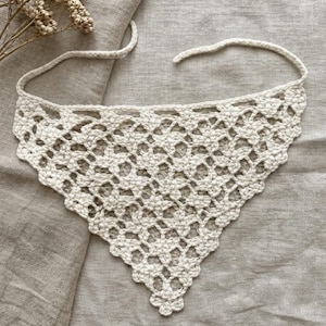 Könnte beinhalten: Cremefarbenes, offen gewebtes Häkel-Bandana mit dreieckiger Form. Das Bandana hat ein zartes, durchbrochenes Muster und lange Bänder zum Befestigen um den Hals. Der Artikel wird auf einer neutralen Leinenoberfläche präsentiert.