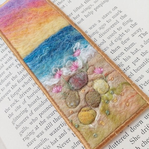 May include: A rectangular needle-felted bookmark featuring a landscape scene with a yellow and orange sky, a blue ocean with white foam, and a sandy beach with embroidered pink flowers, textured pebbles, and green accents, finished with a gold border.