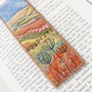 May include: A rectangular fabric bookmark featuring a needle-felted landscape scene with rolling hills in shades of blue, beige, and green, accented by embroidered black dandelion silhouettes, a pink textured line, and small beads on a light background.