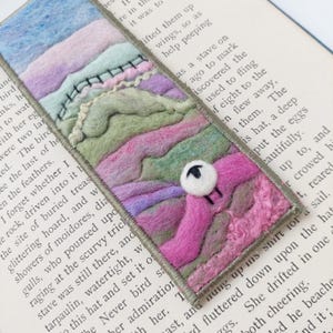 May include: A rectangular needle-felted bookmark featuring layered abstract landscape shapes in shades of blue, lavender, sage green, and magenta. The edges are finished with a grey stitched border, and a small white felted sheep is attached.