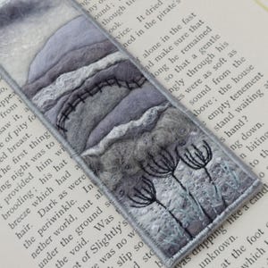 May include: A handcrafted bookmark with a grey landscape design. The bookmark showcases a felted mountain scene with embroidered details, including a fence and delicate floral elements. The edges are neatly finished with a grey border.
