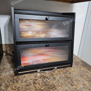 May include: A two-tier black wooden bread box featuring fluted glass panels on the front doors. Each door is equipped with a small circular knob for opening. A silver-toned metal handle is attached to the bottom base of the unit.