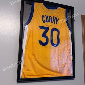May include: Framed yellow basketball jersey with blue accents, featuring the name "CURRY" and the number "30". The jersey is encased in a black frame, ready for display. A signature is visible on the jersey.