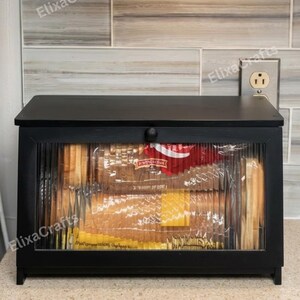 May include: A rectangular black wooden bread box featuring a front-facing door with a fluted glass panel and a small round knob. The interior contains a loaf of sliced bread in a plastic bag with red and yellow packaging visible through the glass.