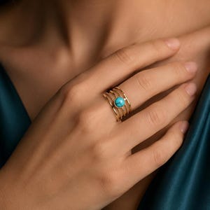Op de afbeelding: Een goudkleurige ring met meerdere banden en een centrale ronde turkoois steen. De ring bestaat uit vier afzonderlijke banden, met twee gladde banden en twee kralenbanden, die allemaal samenkomen bij de zetting van de levendige blauwgroene steen.