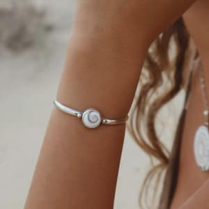 Puede incluir: Una pulsera rígida de metal plateado con un dije circular central que presenta una incrustación de concha en espiral de color blanco y gris. La pulsera tiene un acabado liso y pulido con un diseño sencillo y limpio alrededor de la muñeca.