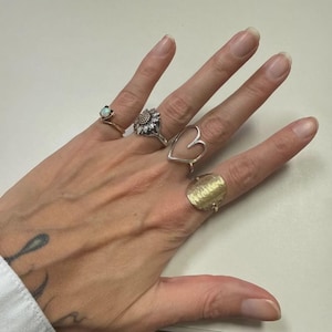 May include: A collection of rings in various designs. Includes a gold ring with an opal, a silver sunflower ring, a silver heart-shaped ring, and a textured gold ring. The rings are displayed on a hand.