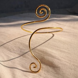 May include: A gold arm cuff featuring a spiral design. The cuff curves around the arm with a circular band and a decorative spiral at the top and bottom. The jewellery is made of a shiny, metallic material.
