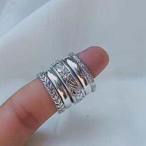 Puede incluir: Un conjunto de anillos de plata con diseños intrincados, incluyendo patrones florales y de hojas. Los anillos están apilados en un dedo, mostrando su detallada artesanía y diversas texturas. Los anillos son de plata.