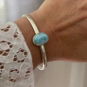 May include: A silver cuff bracelet featuring a smooth, oval-shaped light blue Larimar gemstone set in the center. The metal band has a polished, reflective finish and an open design that wraps around the wrist, showcasing the stone's marbling.