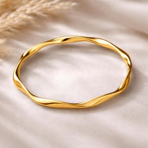 Puede incluir: Un brazalete dorado con un diseño ondulado y orgánico. El brazalete tiene un acabado liso y pulido y está colocado sobre un fondo suave y neutro. El diseño del brazalete es fluido y moderno.