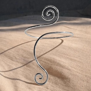 Könnte beinhalten: Ein silberfarbener Armreif mit Spiraldesign. Der Armreif hat ein gebogenes Band, das sich um den Oberarm wickelt, mit einer dekorativen Spirale oben und unten. Der Schmuck wird auf einem neutralfarbenen Hintergrund präsentiert.