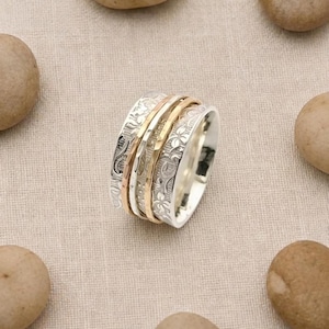 Puede incluir: Un anillo giratorio ancho que presenta una banda central de metal pulido en oro y oro rosa, flanqueada por dos bandas exteriores de plata grabadas con un patrón floral detallado. El anillo está construido con varias capas metálicas.
