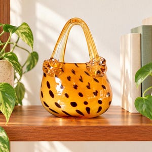 Puede incluir: Una escultura de bolso de mano hecha de vidrio en un tono ámbar cálido, con un patrón repetitivo de manchas de color marrón oscuro. La pieza tiene un asa arqueada transparente y dos pequeños adornos de flores de vidrio con volantes.