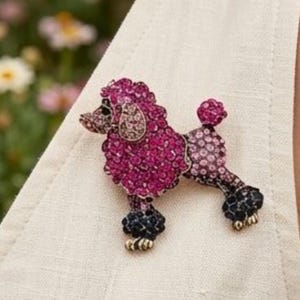 Könnte beinhalten: Eine pudelförmige Brosche, besetzt mit leuchtend pinken, hellrosa und dunkelblauen Kristallen. Die Hundefigur zeigt einen detaillierten Kopf, einen Bommelschwanz und dunkelblaue Kristallakzente an den Pfoten und Ohren auf einem hellen Stoff.