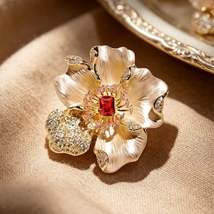 May include: A flower-shaped brooch with a gold-tone frame and creamy white petals. The center features a red gemstone surrounded by pink stones. Accents of clear crystals add sparkle. The brooch is set on a neutral-toned fabric.