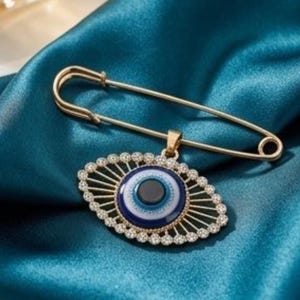 Könnte beinhalten: Eine goldfarbene Sicherheitsnadel-Brosche mit einem zentralen Nazar-Amulett. Der Anhänger ist kreisförmig mit konzentrischen Ringen in Dunkelblau, Hellblau und Weiß, umgeben von einem strahlenförmigen Rahmen, der mit kleinen, klaren Steinen besetzt ist.