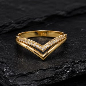 Pode incluir: Um anel dourado em forma de V com um design de banda dupla. A banda superior é incrustada com uma fileira de pequenas pedras claras e brilhantes, enquanto a banda inferior tem um acabamento em ouro liso e polido que se une num ponto central.