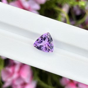 Könnte beinhalten: Ein loser, facettierter Edelstein in dreieckiger Form mit einer leuchtend violetten Farbe. Der Stein weist einen komplexen, symmetrischen Schliff auf, der mehrere lichtreflektierende Oberflächen erzeugt und einen tiefen Lilaton zeigt.