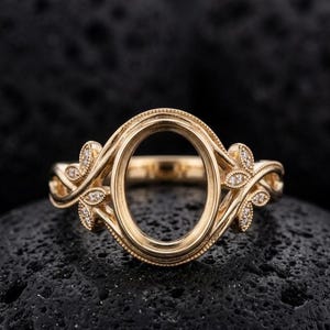 Könnte beinhalten: Ein goldener Ring mit einer leeren, ovalen Fassung in der Mitte, die von einem Perlenrand umgeben ist. Das Band ist auf beiden Seiten mit symmetrischen blattförmigen Akzenten verziert, die mit kleinen, klaren, runden Steinen besetzt sind.