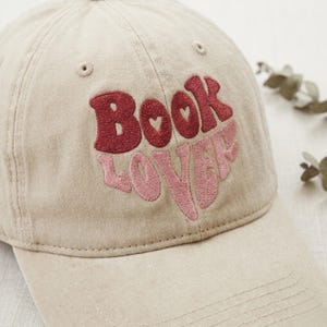 May include: A beige baseball cap with the embroidered text "Book Lover" in red and pink. The cap has a curved brim and a classic six-panel design. The text features heart-shaped details.
