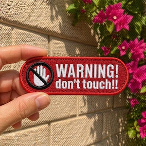 May include: A red embroidered patch with the words "WARNING! don't touch!!" in white. The patch features a black circle with a hand symbol crossed out. The patch is held against a textured background.