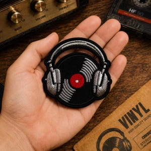 May include: A circular embroidered patch featuring a black vinyl record with a red center label, overlaid with silver-grey headphones. The design uses black, silver, and red thread to create a detailed musical motif suitable for fabric application.