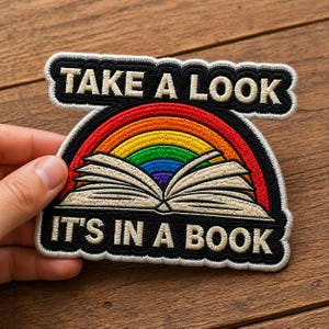 May include: Embroidered patch with the text "TAKE A LOOK IT'S IN A BOOK". The patch features a rainbow arching over an open book. The patch has a black border and white stitching.