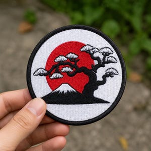 May include: A circular embroidered patch featuring a black bonsai tree silhouette against a large red sun, with a white mountain peak at the base. The design is enclosed within a thick black border on a white background, creating a high-contrast graphic.