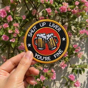 May include: A round, embroidered patch with the text "SHUT UP LIVER YOU'RE FINE" encircling a red background with two beer mugs. The patch has a black border with yellow stitching and two white stars.