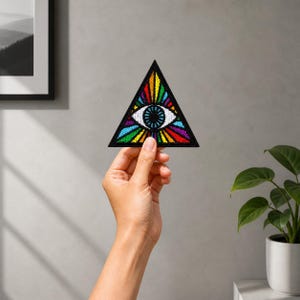 May include: A triangular patch featuring an eye within a triangle, with a rainbow of colors radiating outwards. The patch has a black border and is held up by a hand.